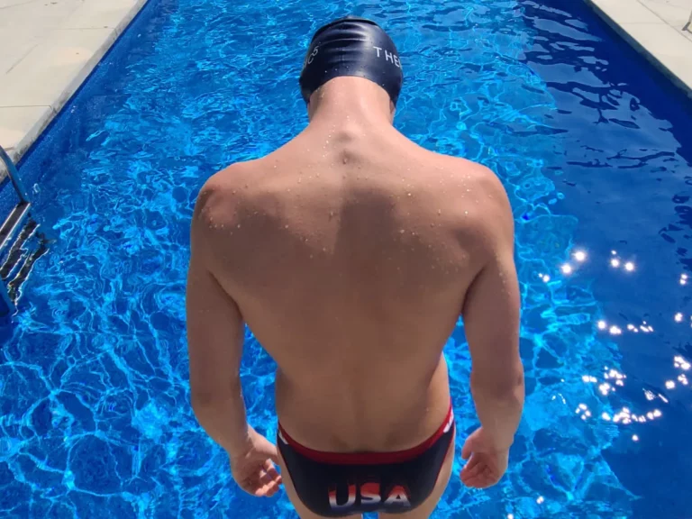 A close-up view from behind of competitive swimmer Alex Aivalotis, standing at the edge of a brilliant blue swimming pool, poised for a dive.